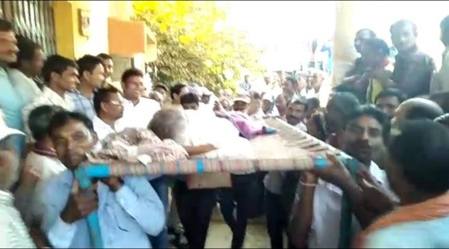 बीमार पति को चारपाई पर लेकर बैंक आई मजबूर पत्नी